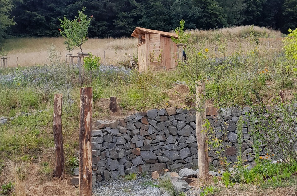 Sitzplatz mit Trockenmauer im Bau, im Hintergrund Luxus-Kompostklo aus Lärchenholz, mit Ausblick übers Tal